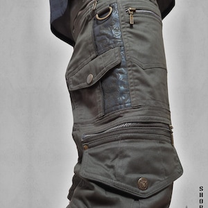 Könnte beinhalten: Olivgrüne Cargoshorts mit mehreren Reißverschlusstaschen, einem D-Ring und einem dunklen Einsatz mit geometrischem Muster. Die Shorts haben metallische Knöpfe und Reißverschlüsse. Der Text lautet SHORTS THETA V2 INDIAN PROJECT.