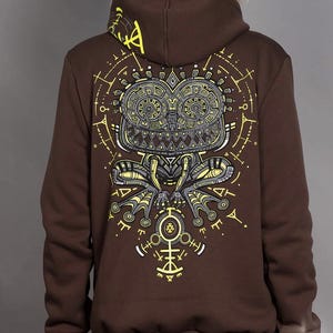 May include: A brown hooded sweatshirt featuring a large, intricate graphic on the back. The design displays a stylized, wide-eyed creature in shades of yellow, grey, and white, surrounded by geometric symbols and lines. The text "plazma lab." is visible.