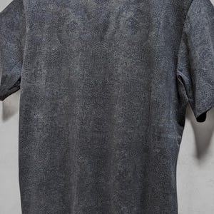 Puede incluir: Una camiseta gris oscura con un patr&oacute;n geom&eacute;trico blanco por toda la superficie. La camiseta tiene cuello redondo y mangas cortas. El texto "INDIAN PROJECT" est&aacute; impreso en la parte superior de la camiseta.