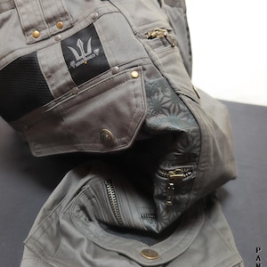 May include: Grey cargo trousers featuring multiple pockets, brass-toned metal rivets, and zips. A black patch displays the text INDIAN PROJECT with a trident logo. A dark panel with a floral pattern is visible near the zip. The text XI Stars INDIAN PROJECT is printed in the corner.
