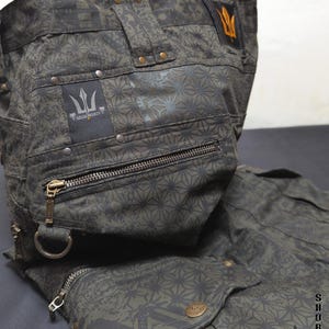 May include: Dark olive green shorts featuring a repeating geometric star pattern, multiple zippered pockets, metal rivets, and a D-ring. A black patch displays the text INDIAN PROJECT with a trident logo. The text THETA V2 INDIAN PROJECT is visible.