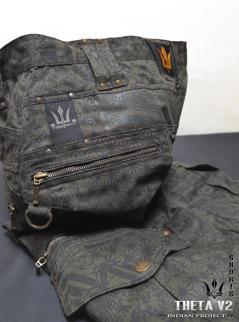 May include: Dark olive green shorts featuring a repeating geometric star pattern, multiple zippered pockets, metal rivets, and a D-ring. A black patch displays the text INDIAN PROJECT with a trident logo. The text THETA V2 INDIAN PROJECT is visible.