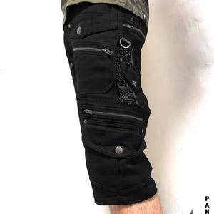 Könnte beinhalten: Schwarze Cargoshorts mit mehreren Reißverschlusstaschen, silberfarbenen Metallbeschlägen und einem dekorativen Einsatz mit geometrischem Muster. Die Shorts verfügen über einen D-Ring und geknöpfte Taschenklappen, wobei der Text THETA STARS INDIAN PROJECT in der Ecke sichtbar ist.