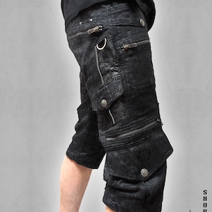 Könnte beinhalten: Schwarze Cargoshorts mit einem dezenten geometrischen Muster, mehreren Reißverschlusstaschen und Metallbeschlägen. Die Shorts verfügen über seitliche Cargotaschen mit Druckknopfverschlüssen und einen D-Ring. Der Text lautet SHORTS THETA V2 INDIAN PROJECT.