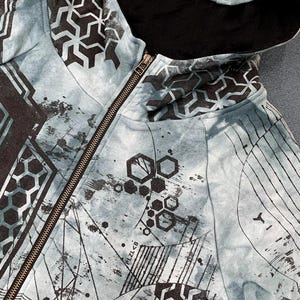 Könnte beinhalten: Ein hellblau-schwarzer Kapuzenpullover mit Reißverschluss, verziert mit einem dunklen geometrischen Muster, sechseckigen Formen und abstrakten Linien. Das Kleidungsstück hat ein schwarzes Innenfutter, einen Reißverschluss und den Aufdruck PLZL<B.