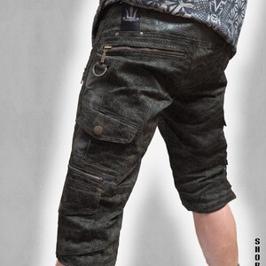 May include: Dark olive green cargo shorts featuring a repeating geometric star pattern, multiple zippered pockets, a metal D-ring, and button-flap cargo pockets. The shorts include a black label with a trident logo and the text THETA V2 INDIAN PROJECT.