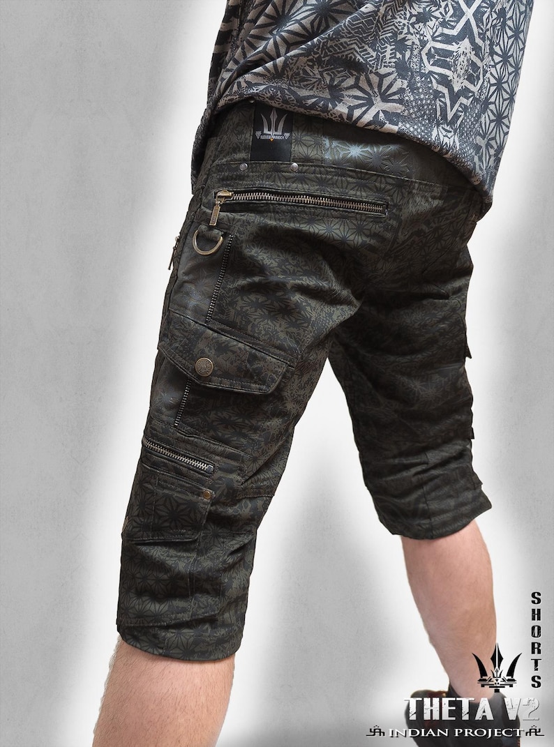 May include: Dark olive green cargo shorts featuring a repeating geometric star pattern, multiple zippered pockets, a metal D-ring, and button-flap cargo pockets. The shorts include a black label with a trident logo and the text THETA V2 INDIAN PROJECT.