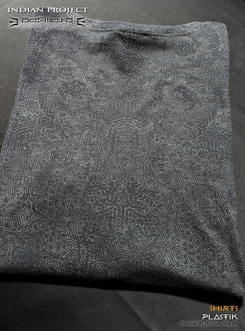 Puede incluir: Una camiseta negra con un patr&oacute;n geom&eacute;trico blanco. La camiseta est&aacute; doblada y sobre una superficie oscura. El texto "SHIRTS PLASTIK" es visible en la esquina inferior derecha de la imagen.