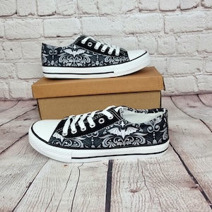 May include: Black and white canvas sneakers with a decorative bat and floral pattern. The shoes have white laces, a white rubber toe cap, and a black stripe along the sole. The sneakers are displayed on a brown cardboard box.