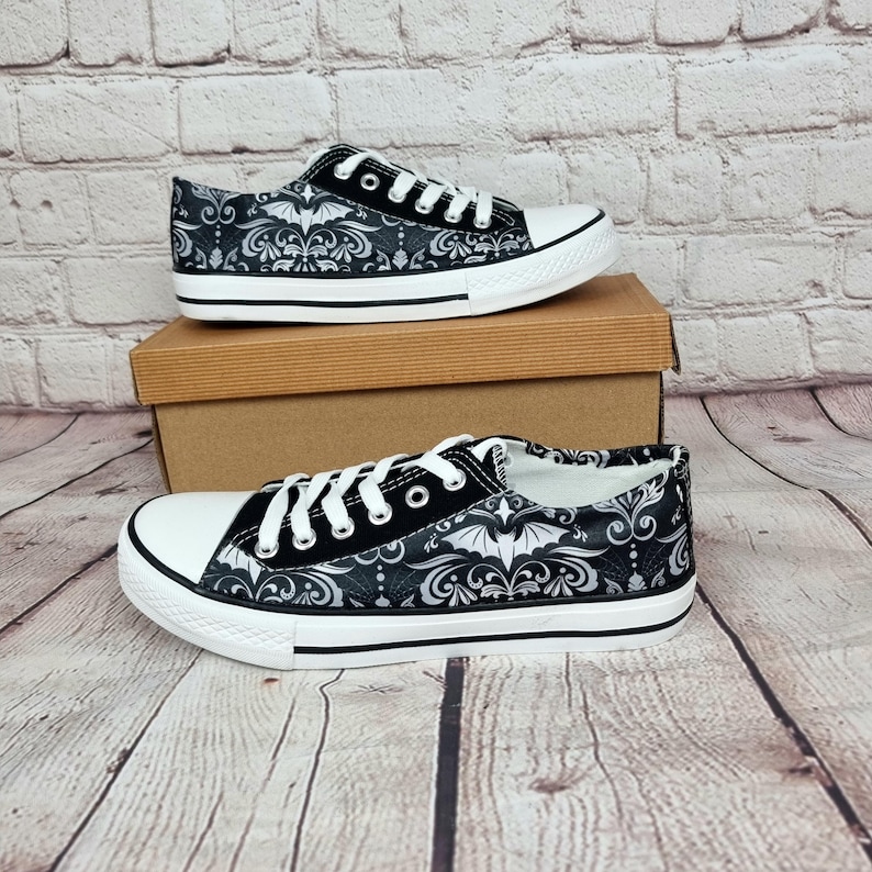 May include: Black and white canvas sneakers with a decorative bat and floral pattern. The shoes have white laces, a white rubber toe cap, and a black stripe along the sole. The sneakers are displayed on a brown cardboard box.