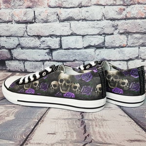May include: A pair of low-top canvas sneakers featuring a dark pattern of human skulls and purple roses. The shoes have white rubber toe caps, white laces, and thick white rubber soles with a thin black stripe running along the side.