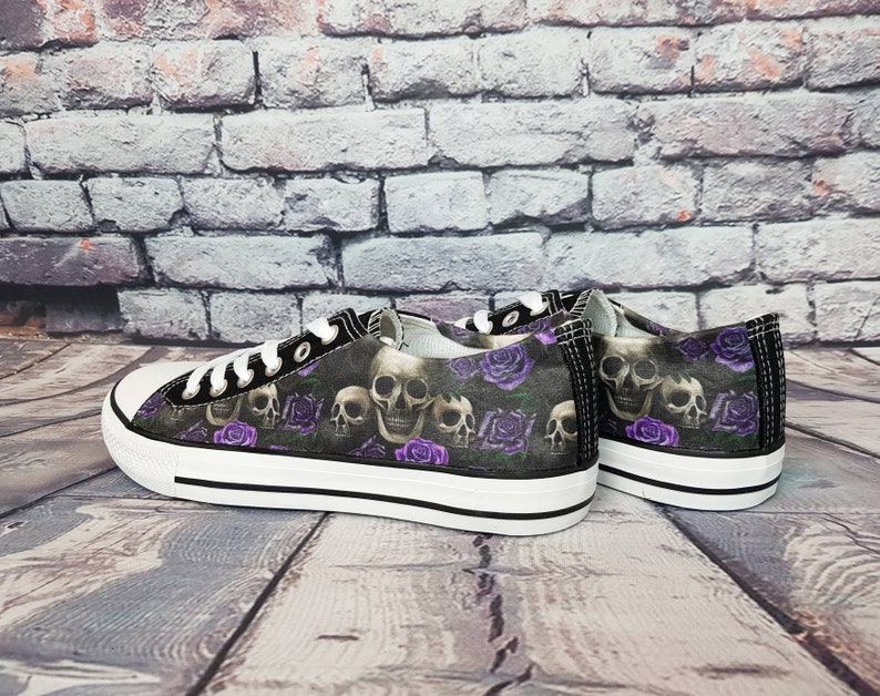 May include: A pair of low-top canvas sneakers featuring a dark pattern of human skulls and purple roses. The shoes have white rubber toe caps, white laces, and thick white rubber soles with a thin black stripe running along the side.