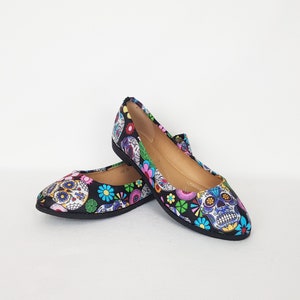 May include: A pair of black ballet flats with a colorful Day of the Dead sugar skull print. The print features skulls with flowers and other decorative elements.