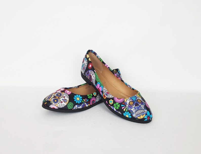 May include: A pair of black ballet flats with a colorful Day of the Dead sugar skull print. The print features skulls with flowers and other decorative elements.