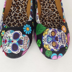 May include: A pair of black canvas flats with a colorful Day of the Dead sugar skull design. The shoes have a leopard print lining.