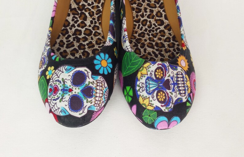 May include: A pair of black canvas flats with a colorful Day of the Dead sugar skull design. The shoes have a leopard print lining.