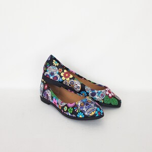 May include: A pair of black flats with a pointed toe. The shoes are decorated with a colorful Day of the Dead sugar skull pattern.