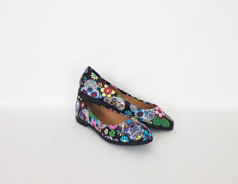 May include: A pair of black flats with a pointed toe. The shoes are decorated with a colorful Day of the Dead sugar skull pattern.