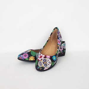 May include: A pair of black flats with a colorful sugar skull and floral print. The shoes have a pointed toe and a low heel.