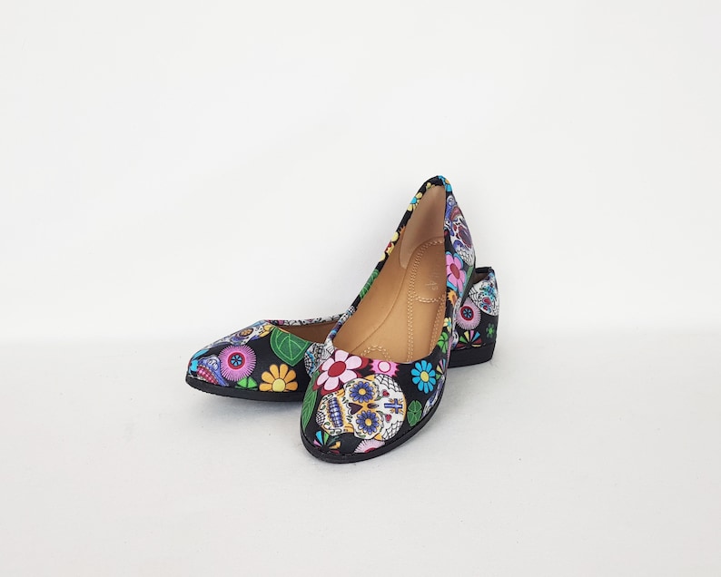 May include: A pair of black flats with a colorful sugar skull and floral print. The shoes have a pointed toe and a low heel.