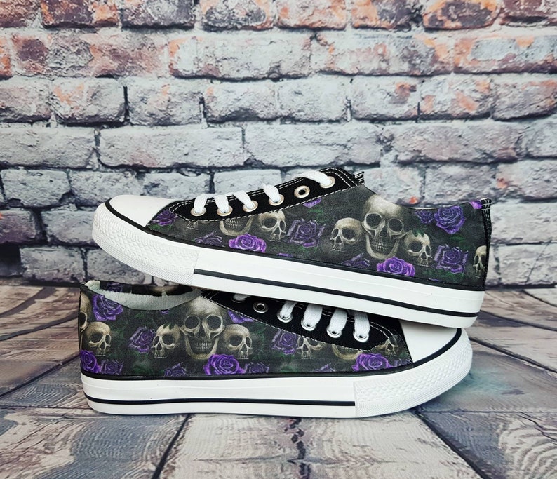 May include: A pair of low-top canvas sneakers featuring a repeating pattern of human skulls and purple roses on a dark background. The shoes have white rubber toe caps, white laces, silver-tone metal eyelets, and white rubber soles with black trim.
