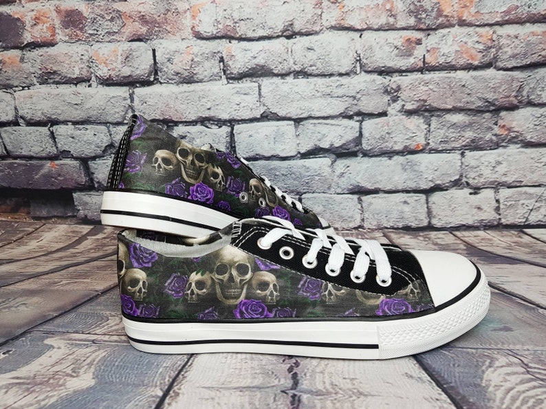 May include: A pair of low-top canvas sneakers featuring a repeating pattern of human skulls and vibrant purple roses on a dark background. The shoes have white rubber toe caps, white laces, and a white rubber sole with a thin black horizontal stripe.