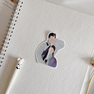 May include: A sticker featuring a cartoon illustration of two people in traditional Korean clothing. The man is holding a sword and the woman is standing behind him. The sticker is on a white gridded notebook.