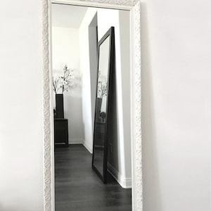 May include: A tall, rectangular white framed mirror with ornate detailing. The mirror is leaning against a white wall, reflecting a dark floor and another mirror. The frame has a decorative pattern around the edges. The mirror is a home decor item.