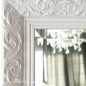 May include: Close-up of a white, ornate mirror frame with detailed floral carvings. The frame is 2.5 inches wide and features a decorative edge. The mirror reflects a chandelier and curtains, adding depth to the image.
