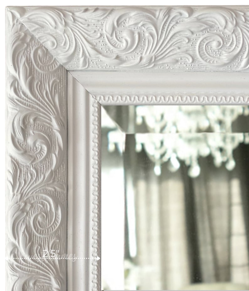 May include: Close-up of a white, ornate mirror frame with detailed floral carvings. The frame is 2.5 inches wide and features a decorative edge. The mirror reflects a chandelier and curtains, adding depth to the image.