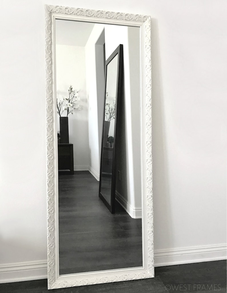 May include: A tall, rectangular white framed mirror with ornate detailing. The mirror is leaning against a white wall, reflecting a dark floor and another mirror. The frame has a decorative pattern around the edges. The mirror is a home decor item.