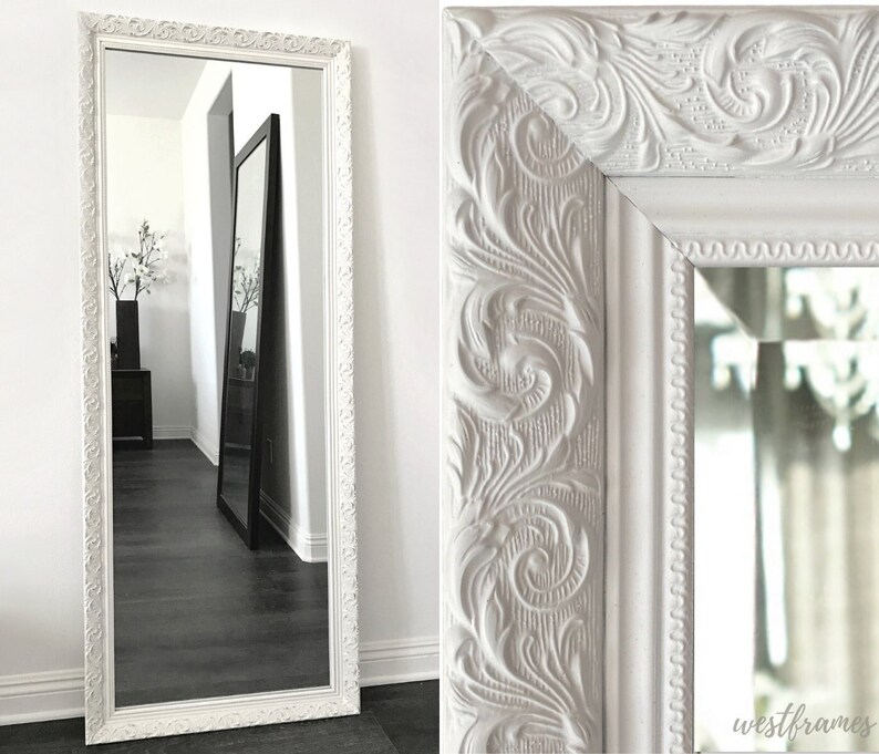 May include: A white, ornate, full-length mirror with a detailed, carved frame. The frame features a scrolling floral design. The mirror is standing on a dark wood floor, reflecting the room. The mirror is from westframes.
