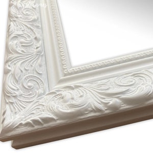 May include: Close-up of a white, ornate picture frame. The frame features detailed, raised floral and scrollwork designs. The frame has a classic, elegant style, with a smooth, matte finish. The West Frames logo is visible in the top left corner.