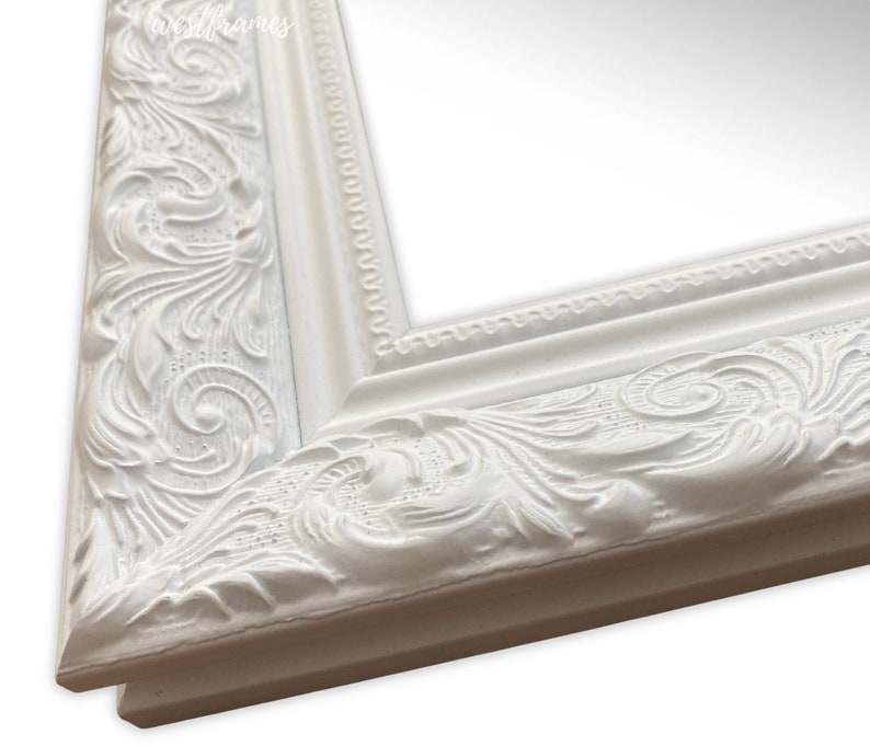 May include: Close-up of a white, ornate picture frame. The frame features detailed, raised floral and scrollwork designs. The frame has a classic, elegant style, with a smooth, matte finish. The West Frames logo is visible in the top left corner.