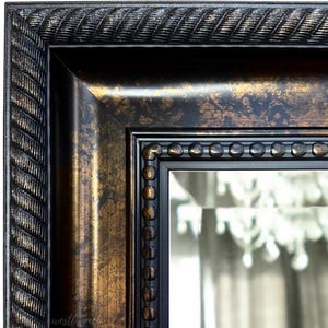 May include: Close-up of a rectangular mirror with a decorative frame. The frame has a dark, distressed finish with gold accents. The outer edge features a rope-like texture, while the inner frame has a beaded design. The mirror reflects a chandelier.