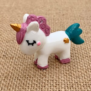 May include: A small white unicorn figurine with pink and purple mane and tail, a gold horn, and a teal tail fin. The figurine has closed eyes and a pink blush on its cheeks.