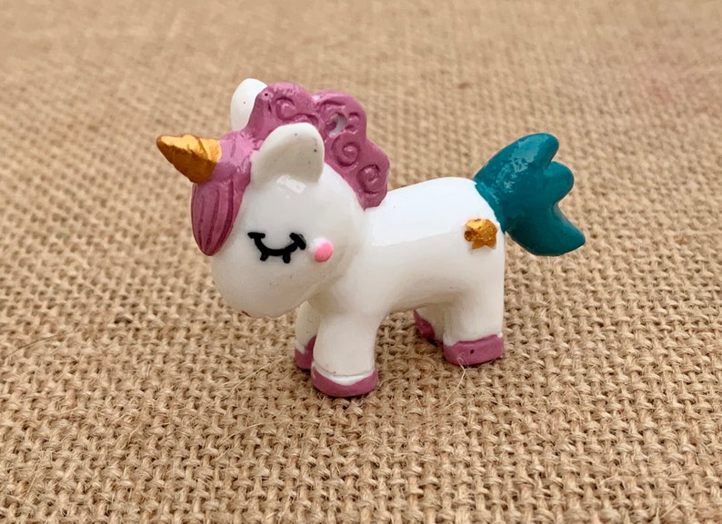 May include: A small white unicorn figurine with pink and purple mane and tail, a gold horn, and a teal tail fin. The figurine has closed eyes and a pink blush on its cheeks.