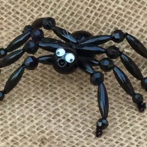 May include: A black beaded spider with googly eyes. The spider has eight legs and a round body. The spider is sitting on a hessian background.