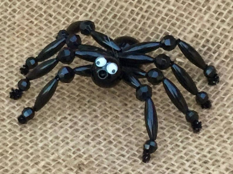 May include: A black beaded spider with googly eyes. The spider has eight legs and a round body. The spider is sitting on a hessian background.