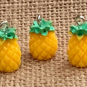 May include: Three yellow pineapple charms with green tops and silver-colored loops for jewelry making. The charms have a textured surface and are arranged on a neutral background.