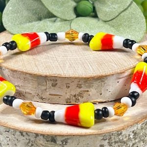 Può includere: Un braccialetto con ciondoli a forma di mais dolce con perline nere, bianche e arancioni. I ciondoli a forma di mais dolce sono gialli, arancioni e bianchi.