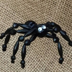 May include: A black beaded spider with googly eyes. The spider has eight legs and a round body. The spider is sitting on a hessian background.