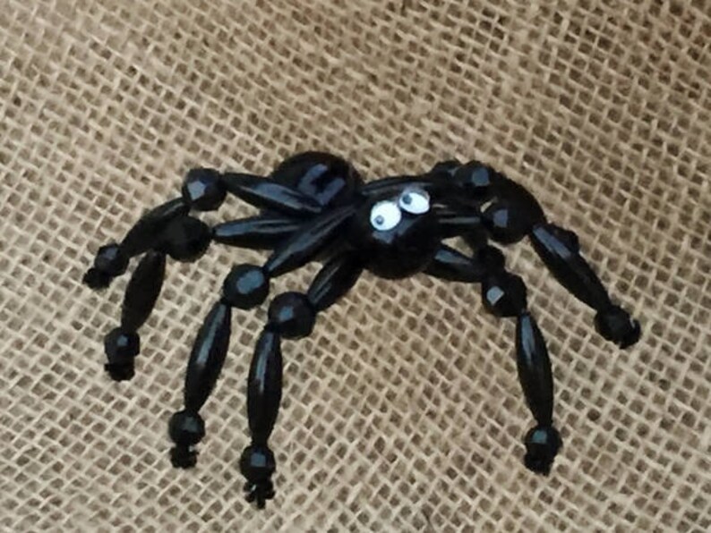 May include: A black beaded spider with googly eyes. The spider has eight legs and a round body. The spider is sitting on a hessian background.
