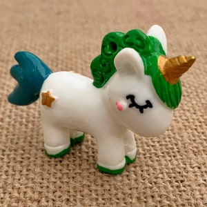 May include: A white unicorn figurine with a green mane and tail, gold horn, and gold star accents. The unicorn is standing on a brown hessian surface.