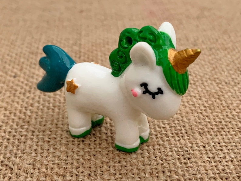 May include: A white unicorn figurine with a green mane and tail, gold horn, and gold star accents. The unicorn is standing on a brown hessian surface.