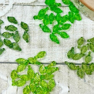 May include: Various translucent green leaf-shaped beads scattered on a light-coloured wooden surface. The beads vary in shades of green. These decorative beads are ideal for crafting and are measured in centimetres.