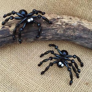 May include: Two black beaded spiders with white eyes. The spiders are crafted with black beads and have a shiny finish. The spiders are sitting on a piece of driftwood.