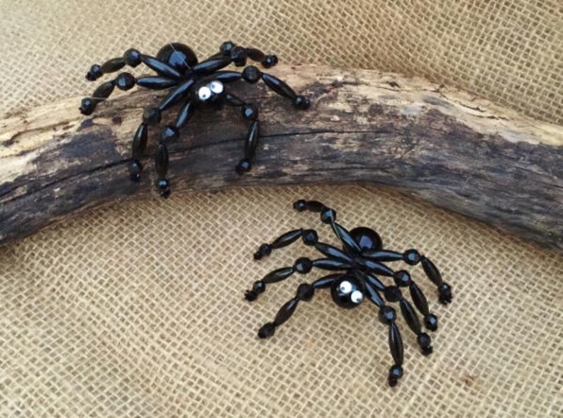 May include: Two black beaded spiders with white eyes. The spiders are crafted with black beads and have a shiny finish. The spiders are sitting on a piece of driftwood.