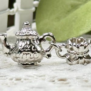 Könnte beinhalten: Ein glänzender silberfarbener Metallanhänger in Form einer Miniatur-Teekanne mit der Prägung "tea time" an der Seite, der durch einen kleinen Biegering mit einer passenden Miniatur-Teetasse mit gewelltem Rand und texturiertem Boden verbunden ist.