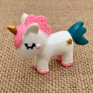 May include: A small white unicorn figurine with pink hair, a gold horn, and a teal tail. The unicorn has closed eyes and a gold star on its side.